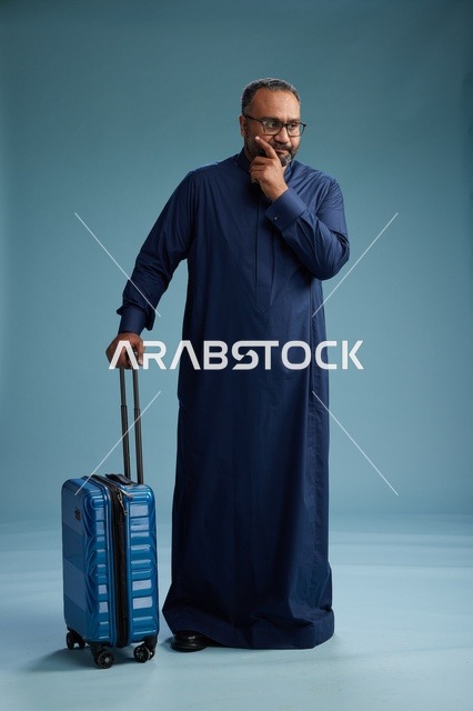 بورتيه لرجل  عربي سعودي  خليجي بثوب أزرق واقف مع حقيبة سفر, الاستعداد للسفر  والسياحه, الجسم بالطول الكامل, خلفيه زرقاء