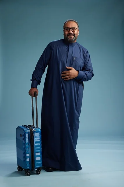 A portrait of a Saudi Arabian Gulf man in a blue thobe standing with a suitcase, ready for travel and tourism, full-body shot, blue background.