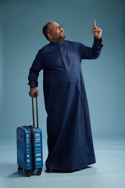 A portrait of a Saudi Arabian Gulf man in a blue thobe standing with a suitcase, ready for travel and tourism, full-body shot, blue background.