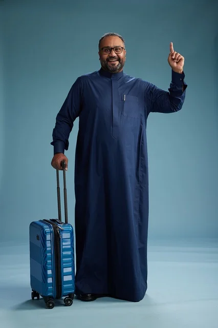 A portrait of a Saudi Arabian Gulf man in a blue thobe standing with a suitcase, ready for travel and tourism, full-body shot, blue background.