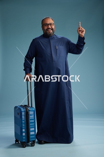 بورتيه لرجل  عربي سعودي  خليجي بثوب أزرق واقف مع حقيبة سفر, الاستعداد للسفر  والسياحه, الجسم بالطول الكامل, خلفيه زرقاء
