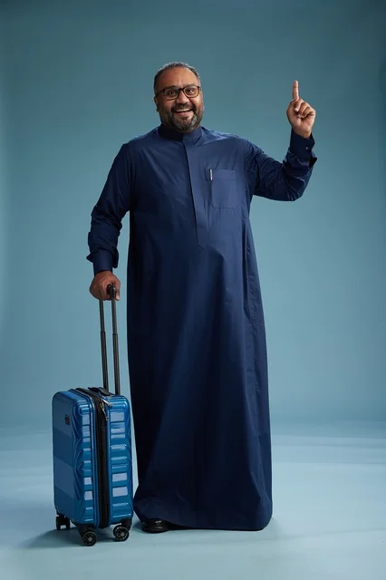 A portrait of a Saudi Arabian Gulf man in a blue thobe standing with a suitcase, ready for travel and tourism, full-body shot, blue background.