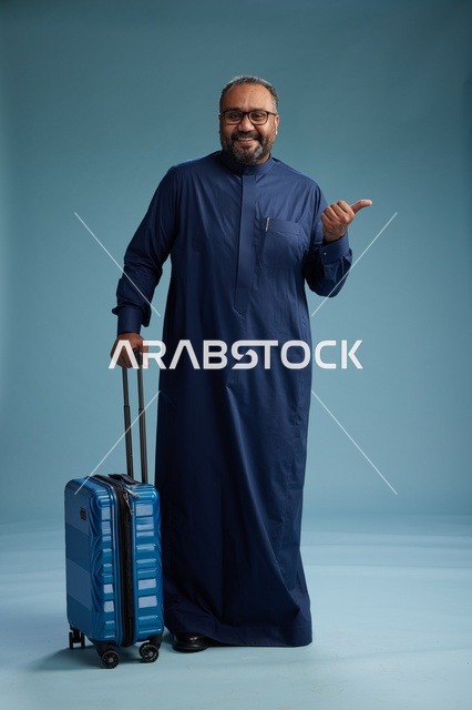 A portrait of a Saudi Arabian Gulf man in a blue thobe standing with a suitcase, ready for travel and tourism, full-body shot, blue background.