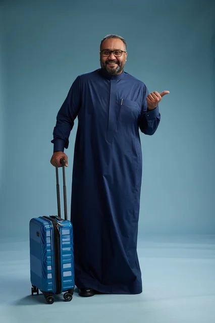 A portrait of a Saudi Arabian Gulf man in a blue thobe standing with a suitcase, ready for travel and tourism, full-body shot, blue background.