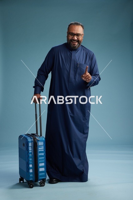 بورتيه لرجل  عربي سعودي  خليجي بثوب أزرق واقف مع حقيبة سفر, الاستعداد للسفر  والسياحه, الجسم بالطول الكامل, خلفيه زرقاء