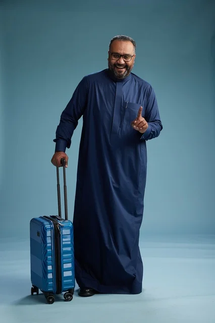 A portrait of a Saudi Arabian Gulf man in a blue thobe standing with a suitcase, ready for travel and tourism, full-body shot, blue background.