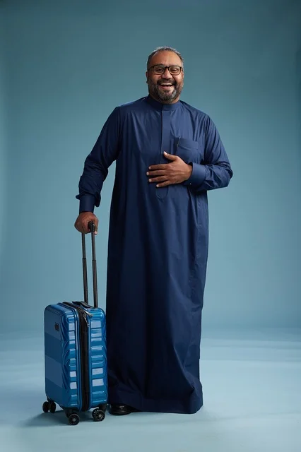 A portrait of a Saudi Arabian Gulf man in a blue thobe standing with a suitcase, ready for travel and tourism, full-body shot, blue background.