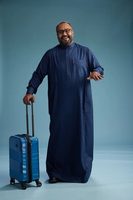 A portrait of a Saudi Arabian Gulf man in a blue thobe standing with a suitcase, ready for travel and tourism, full-body shot, blue background.
