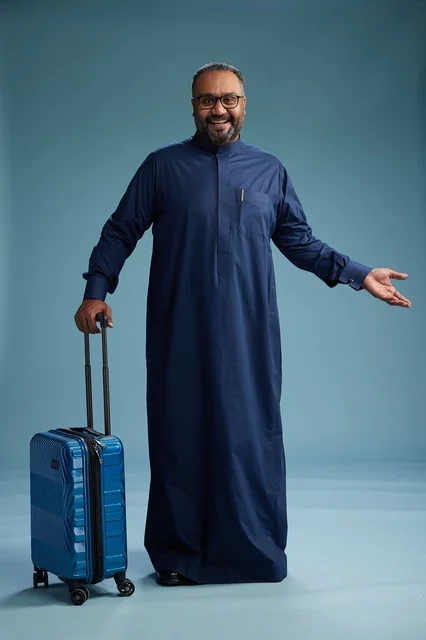 A portrait of a Saudi Arabian Gulf man in a blue thobe standing with a suitcase, ready for travel and tourism, full-body shot, blue background.