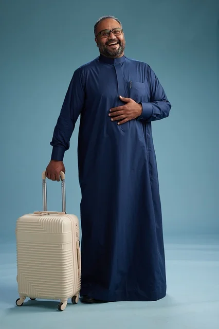 A portrait of a Saudi Arabian Gulf man in a blue thobe standing with a suitcase, ready for travel and tourism, full-body shot, blue background.