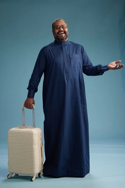 A portrait of a Saudi Arabian Gulf man in a blue thobe standing with a suitcase, ready for travel and tourism, full-body shot, blue background.