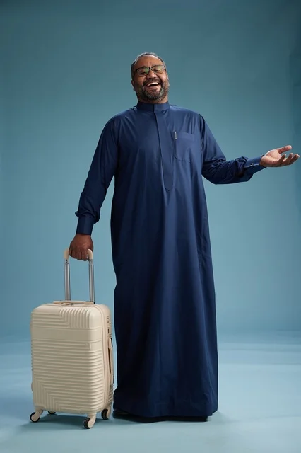 A portrait of a Saudi Arabian Gulf man in a blue thobe standing with a suitcase, ready for travel and tourism, full-body shot, blue background.