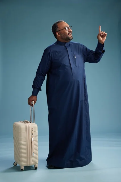 A portrait of a Saudi Arabian Gulf man in a blue thobe standing with a suitcase, ready for travel and tourism, full-body shot, blue background. A portrait of a Saudi Arabian Gulf man in a blue thobe standing with a suitcase, ready for travel and tourism, full-body shot, blue background.