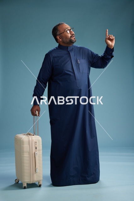 بورتيه لرجل  عربي سعودي  خليجي بثوب أزرق واقف مع حقيبة سفر, الاستعداد للسفر  والسياحه, الجسم بالطول الكامل, خلفيه زرقاء