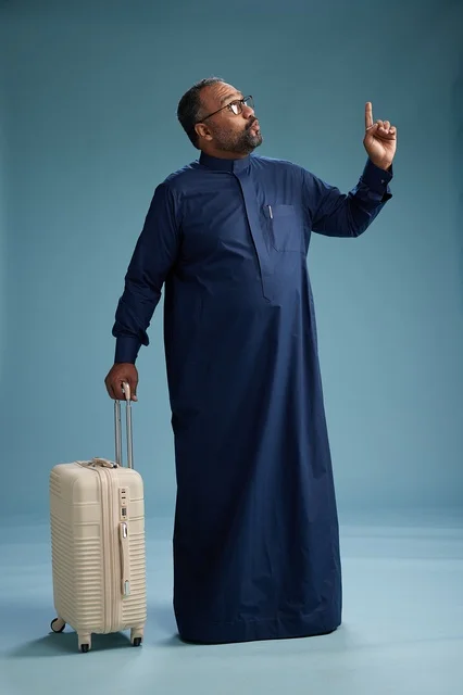 A portrait of a Saudi Arabian Gulf man in a blue thobe standing with a suitcase, ready for travel and tourism, full-body shot, blue background.