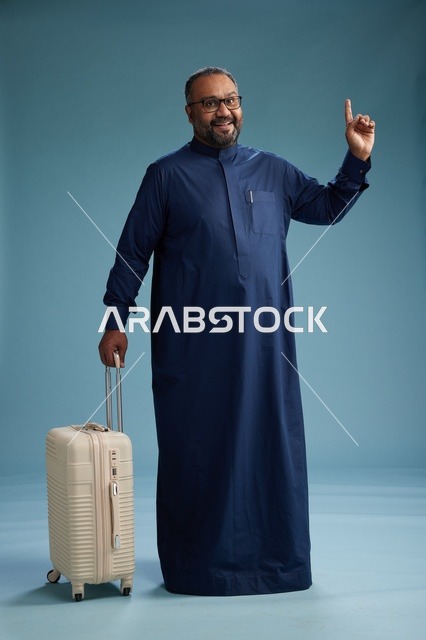بورتيه لرجل  عربي سعودي  خليجي بثوب أزرق واقف مع حقيبة سفر, الاستعداد للسفر  والسياحه, الجسم بالطول الكامل, خلفيه زرقاء