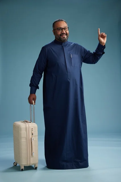 A portrait of a Saudi Arabian Gulf man in a blue thobe standing with a suitcase, ready for travel and tourism, full-body shot, blue background.