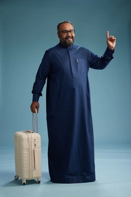 A portrait of a Saudi Arabian Gulf man in a blue thobe standing with a suitcase, ready for travel and tourism, full-body shot, blue background.