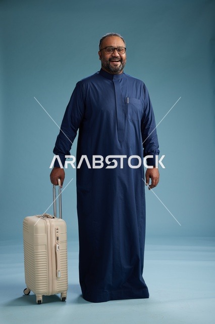 بورتيه لرجل  عربي سعودي  خليجي بثوب أزرق واقف مع حقيبة سفر, الاستعداد للسفر  والسياحه, الجسم بالطول الكامل, خلفيه زرقاء