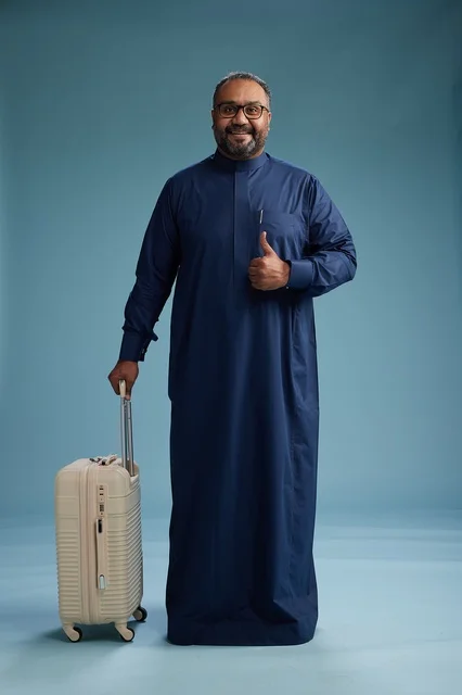 A portrait of a Saudi Arabian Gulf man in a blue thobe standing with a suitcase, ready for travel and tourism, full-body shot, blue background. A portrait of a Saudi Arabian Gulf man in a blue thobe standing with a suitcase, ready for travel and tourism, full-body shot, blue background.
