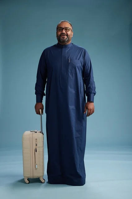 A portrait of a Saudi Arabian Gulf man in a blue thobe standing with a suitcase, ready for travel and tourism, full-body shot, blue background.