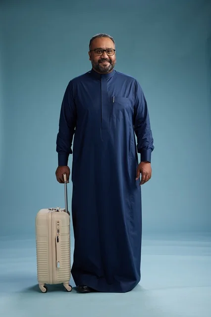 A portrait of a Saudi Arabian Gulf man in a blue thobe standing with a suitcase, ready for travel and tourism, full-body shot, blue background.
