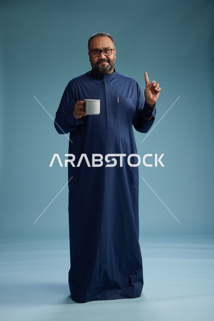 بورتريه لرجل عربي سعودي  خليجي بثوب أزرق يحمل كوب قهوة ويبتسم, الاستمتاع بالمشروب الساخن, الجسم بالطول الكامل, خلفيه زرقاء