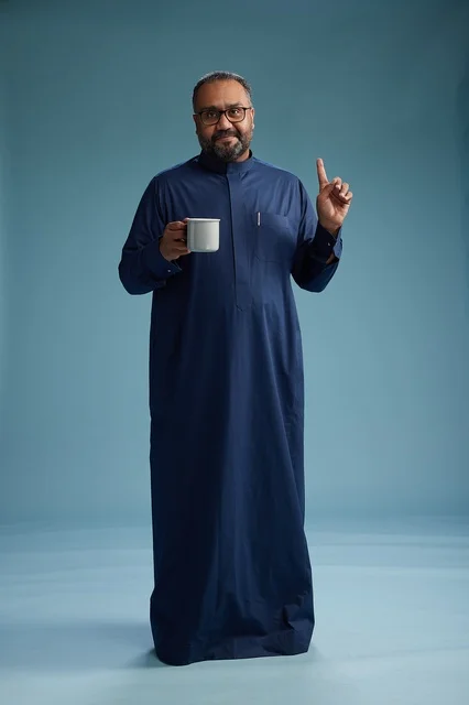 A portrait of a Saudi Arabian Gulf man in a blue thobe holding a cup of coffee and smiling, enjoying the hot drink, full-body shot, with a blue background. A portrait of a Saudi Arabian Gulf man in a blue thobe holding a cup of coffee and smiling, enjoying the hot drink, full-body shot, with a blue background.