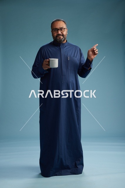 بورتريه لرجل عربي سعودي  خليجي بثوب أزرق يحمل كوب قهوة ويبتسم, الاستمتاع بالمشروب الساخن, الجسم بالطول الكامل, خلفيه زرقاء