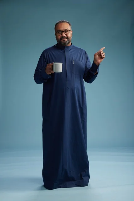 A portrait of a Saudi Arabian Gulf man in a blue thobe holding a cup of coffee and smiling, enjoying the hot drink, full-body shot, with a blue background.
