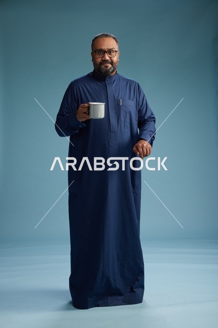 بورتريه لرجل عربي سعودي  خليجي بثوب أزرق يحمل كوب قهوة ويبتسم, الاستمتاع بالمشروب الساخن, الجسم بالطول الكامل, خلفيه زرقاء