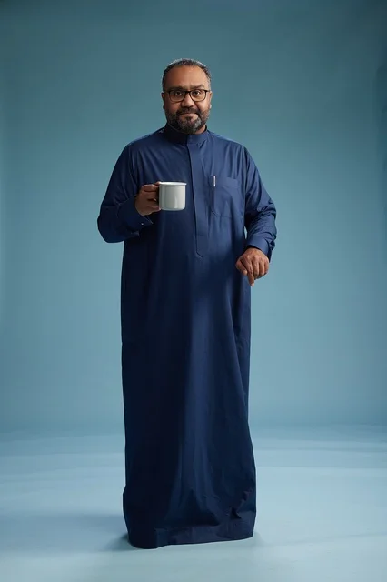 A portrait of a Saudi Arabian Gulf man in a blue thobe holding a cup of coffee and smiling, enjoying the hot drink, full-body shot, with a blue background.