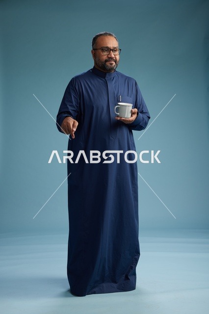 بورتريه لرجل عربي سعودي  خليجي بثوب أزرق يحمل كوب قهوة ويبتسم, الاستمتاع بالمشروب الساخن, الجسم بالطول الكامل, خلفيه زرقاء