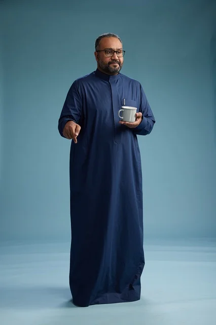 A portrait of a Saudi Arabian Gulf man in a blue thobe holding a cup of coffee and smiling, enjoying the hot drink, full-body shot, with a blue background.