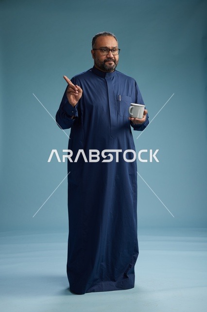بورتريه لرجل عربي سعودي  خليجي بثوب أزرق يحمل كوب قهوة ويبتسم, الاستمتاع بالمشروب الساخن, الجسم بالطول الكامل, خلفيه زرقاء