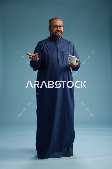 بورتريه لرجل عربي سعودي  خليجي بثوب أزرق يحمل كوب قهوة ويبتسم, الاستمتاع بالمشروب الساخن, الجسم بالطول الكامل, خلفيه زرقاء