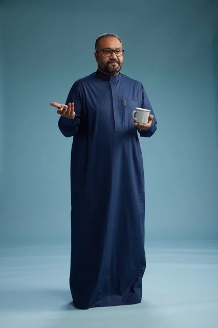 A portrait of a Saudi Arabian Gulf man in a blue thobe holding a cup of coffee and smiling, enjoying the hot drink, full-body shot, with a blue background.