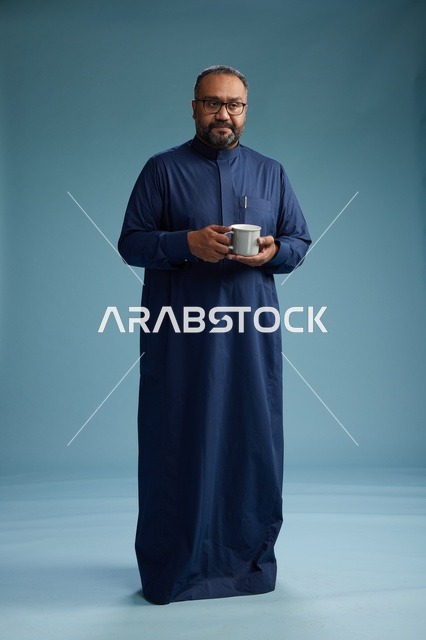 بورتريه لرجل عربي سعودي  خليجي بثوب أزرق يحمل كوب قهوة ويبتسم, الاستمتاع بالمشروب الساخن, الجسم بالطول الكامل, خلفيه زرقاء