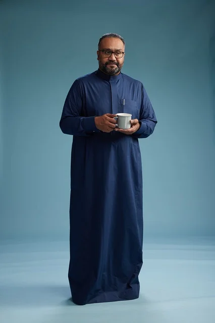 A portrait of a Saudi Arabian Gulf man in a blue thobe holding a cup of coffee and smiling, enjoying the hot drink, full-body shot, with a blue background.