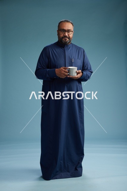 بورتريه لرجل عربي سعودي  خليجي بثوب أزرق يحمل كوب قهوة ويبتسم, الاستمتاع بالمشروب الساخن, الجسم بالطول الكامل, خلفيه زرقاء