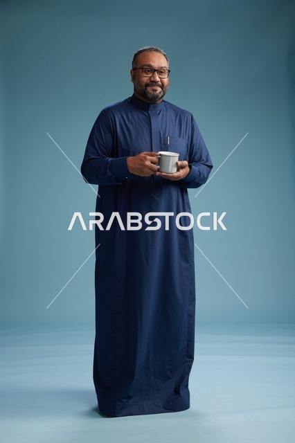 بورتريه لرجل عربي سعودي  خليجي بثوب أزرق يحمل كوب قهوة ويبتسم, الاستمتاع بالمشروب الساخن, الجسم بالطول الكامل, خلفيه زرقاء