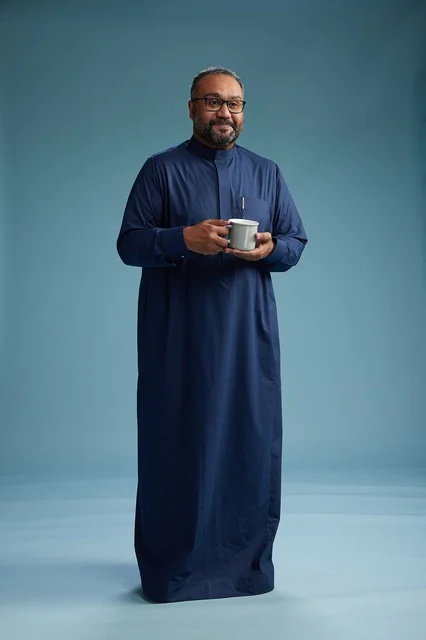 بورتريه لرجل عربي سعودي  خليجي بثوب أزرق يحمل كوب قهوة ويبتسم, الاستمتاع بالمشروب الساخن, الجسم بالطول الكامل, خلفيه زرقاء