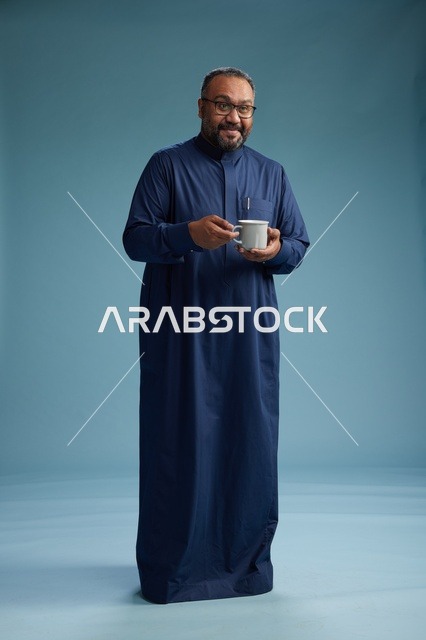بورتريه لرجل عربي سعودي  خليجي بثوب أزرق يحمل كوب قهوة ويبتسم, الاستمتاع بالمشروب الساخن, الجسم بالطول الكامل, خلفيه زرقاء