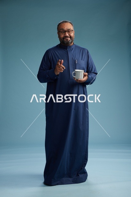 بورتريه لرجل عربي سعودي  خليجي بثوب أزرق يحمل كوب قهوة ويبتسم, الاستمتاع بالمشروب الساخن, الجسم بالطول الكامل, خلفيه زرقاء