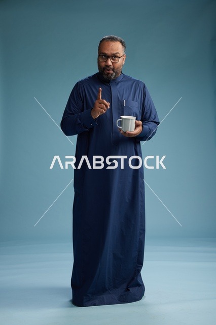 بورتريه لرجل عربي سعودي  خليجي بثوب أزرق يحمل كوب قهوة ويبتسم, الاستمتاع بالمشروب الساخن, الجسم بالطول الكامل, خلفيه زرقاء