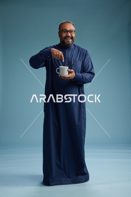 بورتريه لرجل عربي سعودي  خليجي بثوب أزرق يحمل كوب قهوة ويبتسم, الاستمتاع بالمشروب الساخن, الجسم بالطول الكامل, خلفيه زرقاء