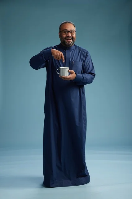 بورتريه لرجل عربي سعودي  خليجي بثوب أزرق يحمل كوب قهوة ويبتسم, الاستمتاع بالمشروب الساخن, الجسم بالطول الكامل, خلفيه زرقاء