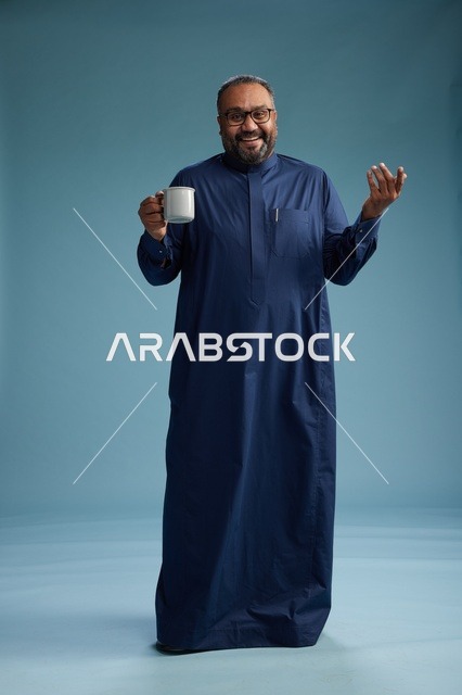 بورتريه لرجل عربي سعودي  خليجي بثوب أزرق يحمل كوب قهوة ويبتسم, الاستمتاع بالمشروب الساخن, الجسم بالطول الكامل, خلفيه زرقاء
