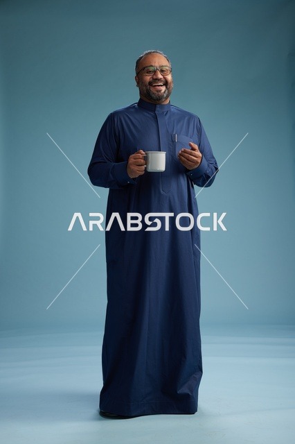 بورتريه لرجل عربي سعودي  خليجي بثوب أزرق يحمل كوب قهوة ويبتسم, الاستمتاع بالمشروب الساخن, الجسم بالطول الكامل, خلفيه زرقاء