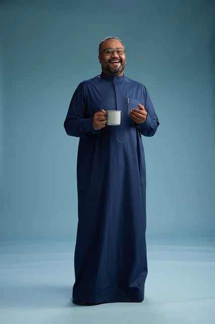 بورتريه لرجل عربي سعودي  خليجي بثوب أزرق يحمل كوب قهوة ويبتسم, الاستمتاع بالمشروب الساخن, الجسم بالطول الكامل, خلفيه زرقاء
